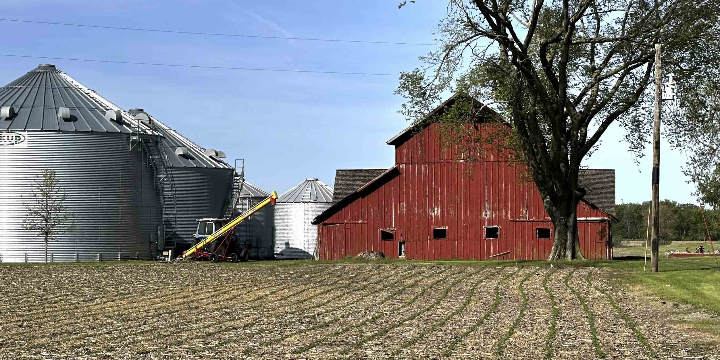 A red barn and grain bins beside a freshly planted Iowa field under a blue sky.