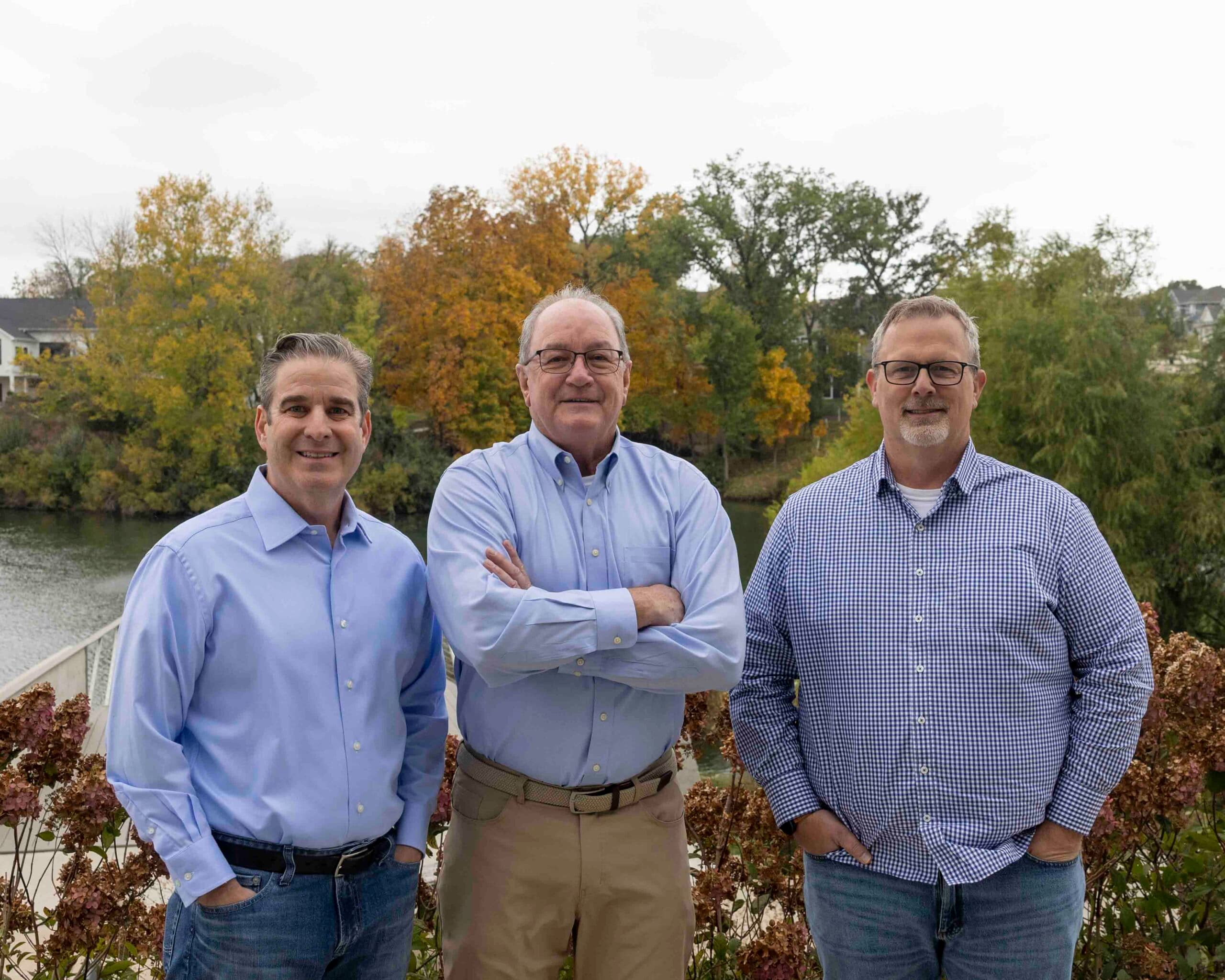 Three business brokers standing together outdoors beside a tree-lined river on an overcast autumn day.